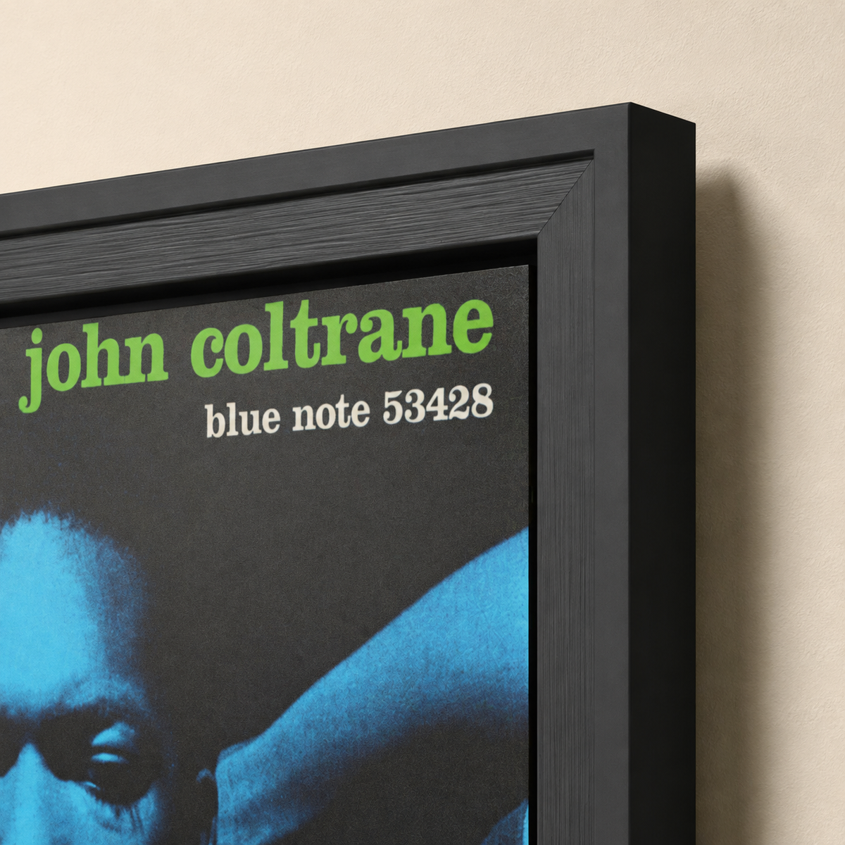 A framed John Coltrane poster shown at an angle against a beige wall. The black frame contains a blue-toned portrait and green and white text reading 'john coltrane' and 'blue note 53428'.