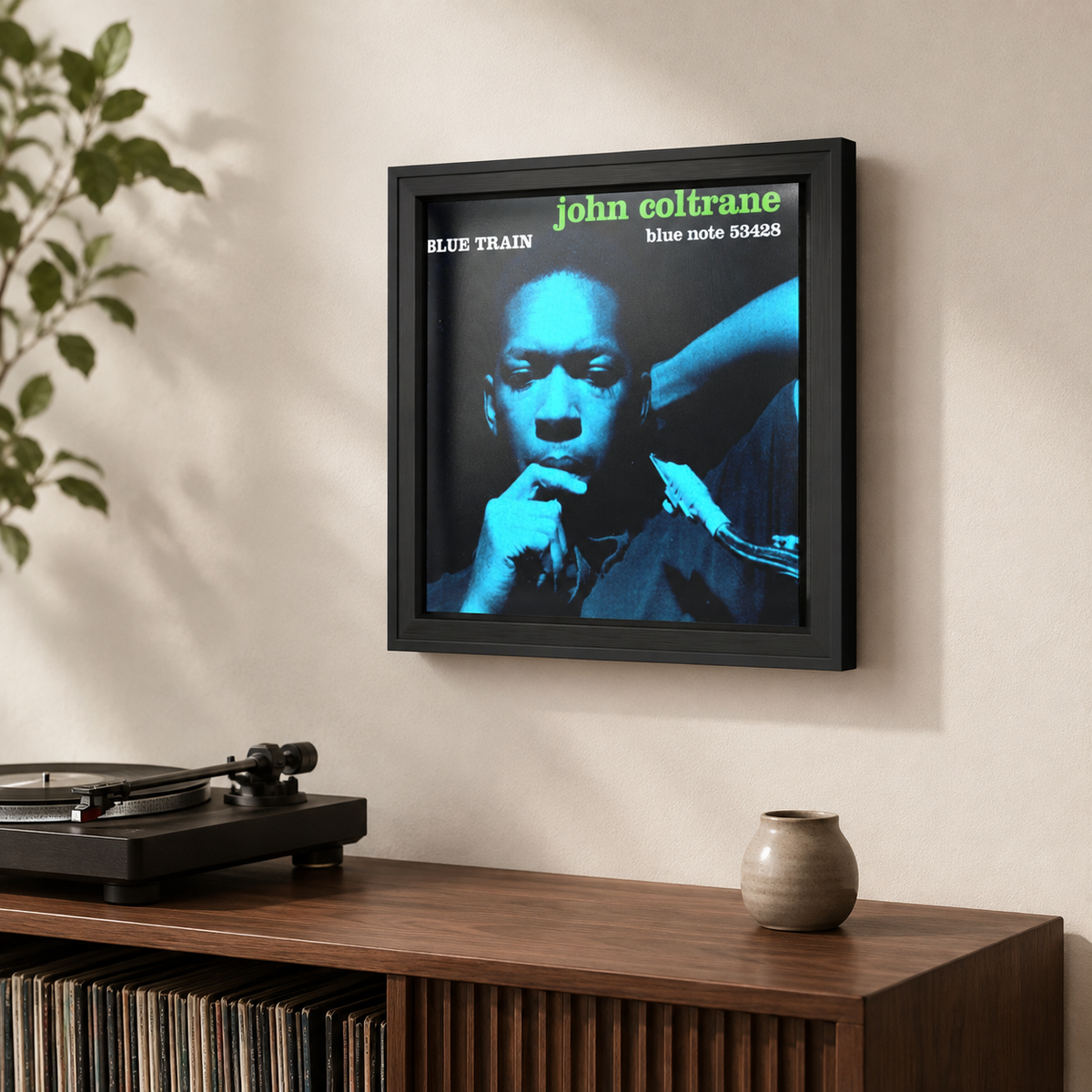 A framed square wall art print of John Coltrane's 'Blue Train' album cover hanging on a beige wall above a wooden record cabinet, with a turntable and small vase nearby.