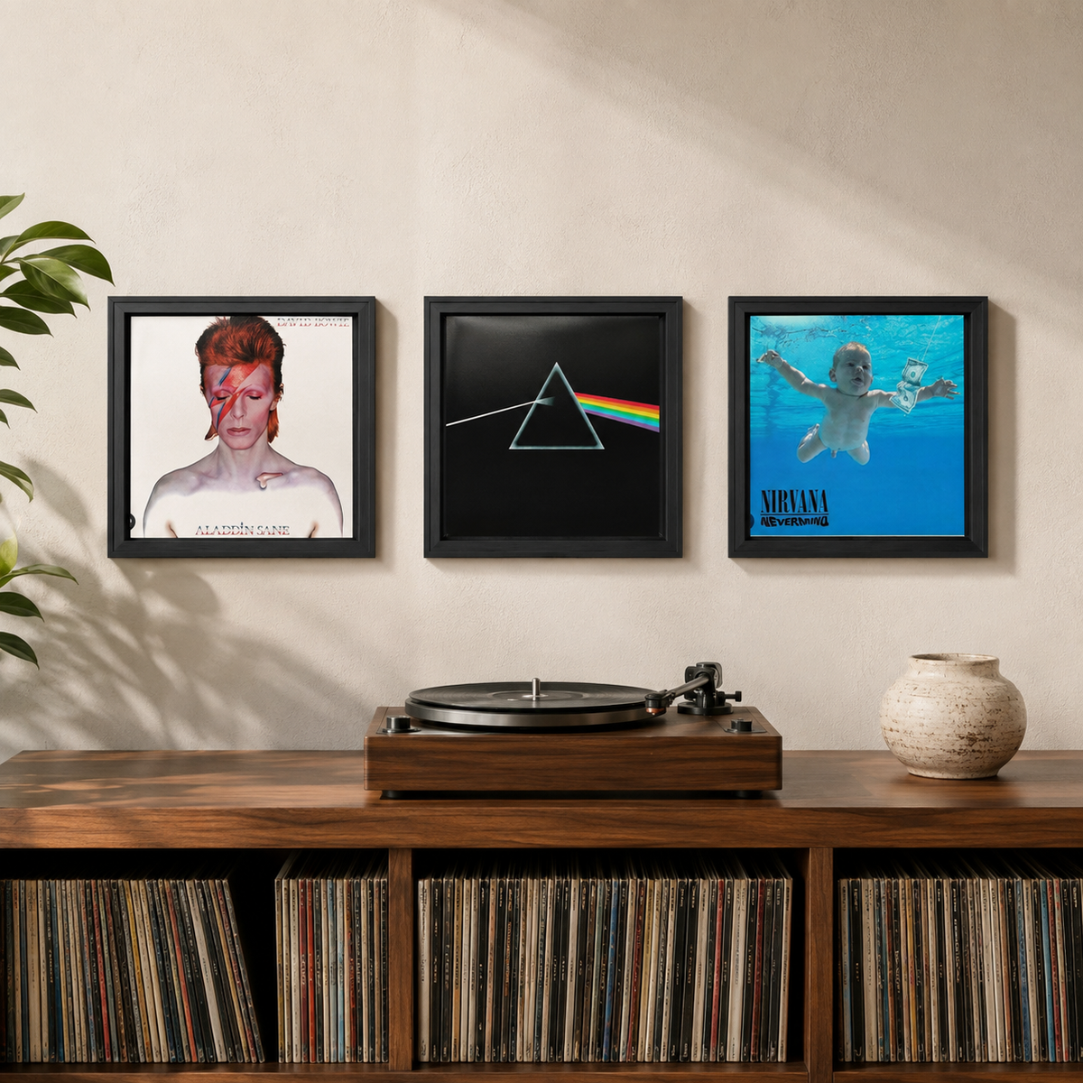 Three framed music-themed wall art prints hang above a wooden record cabinet in a styled room. The left print shows David Bowie with a red eye patch and the text 'Alhambra'; the center print shows the Pink Floyd prism logo on a black background; the right print shows Nirvana on blue album art with the text 'Mental Health'.